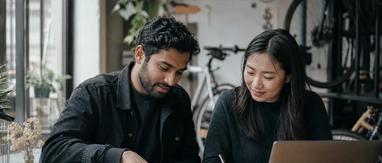 The owner of a small online bike-parts shop points to a page in a printed handbook while briefing a freelance developer at a café table.