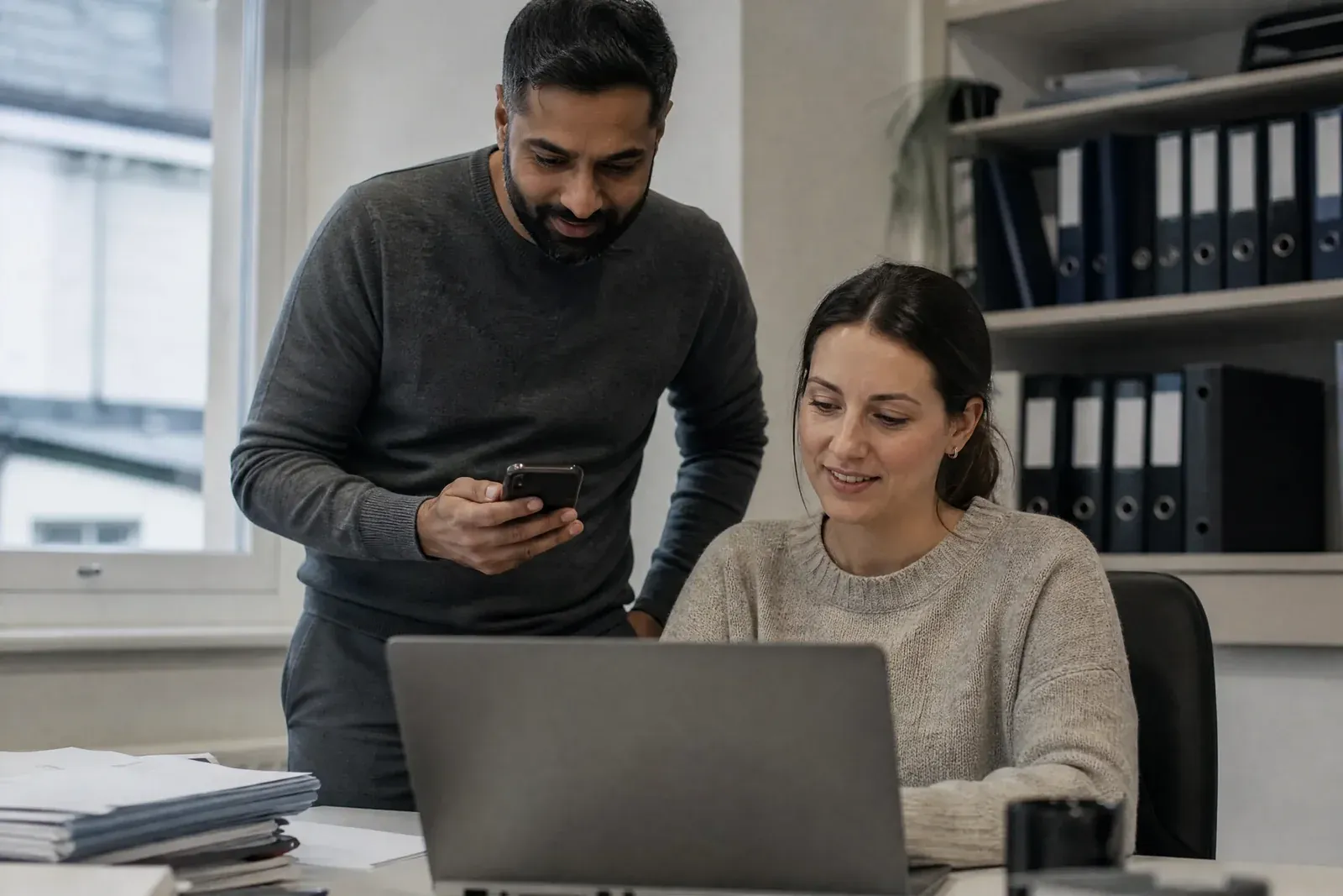 Two people test a website in a small office: one submits a test enquiry on a phone while the other watches a debugging view on a laptop.