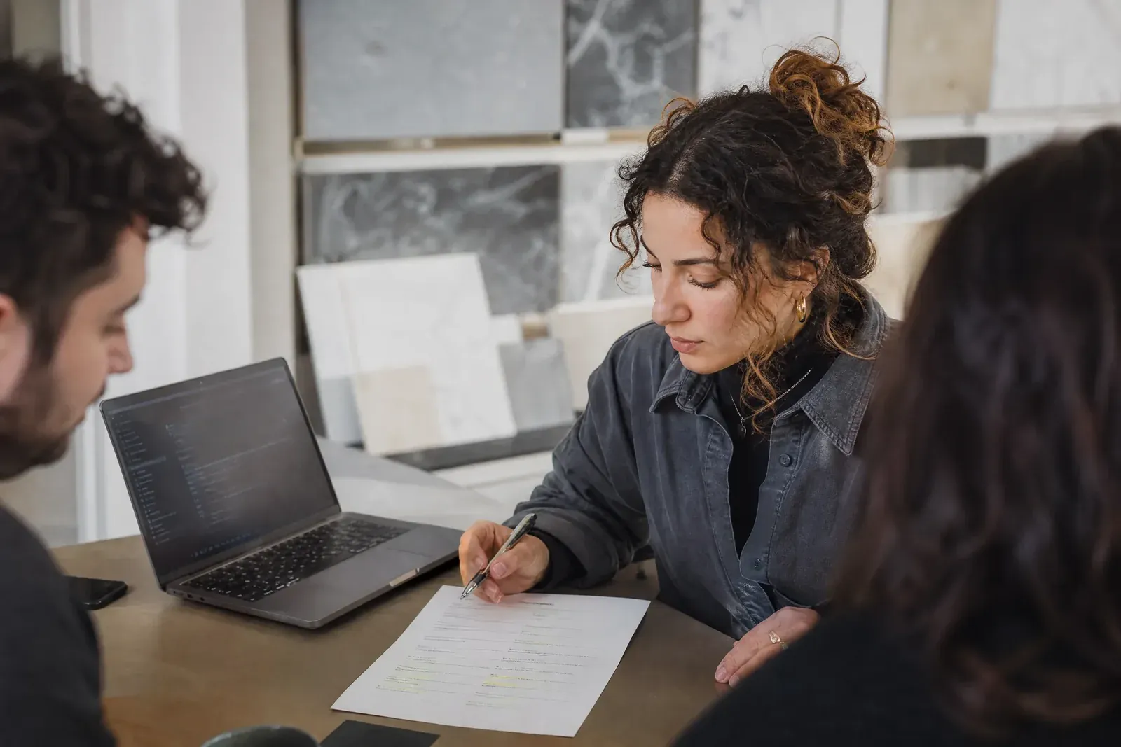The owner of a small service business sits in a showroom office with a laptop open to an AI chat and a printed list of search terms with some lines highlighted.